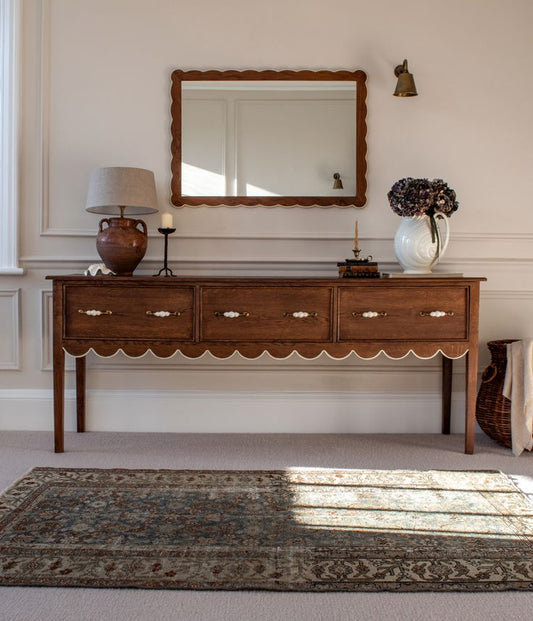 Vintage terracotta lamp with looped handles and neutral linen shade, styled on a scalloped wooden sideboard beneath a carved mirror in a warm-toned hallway.