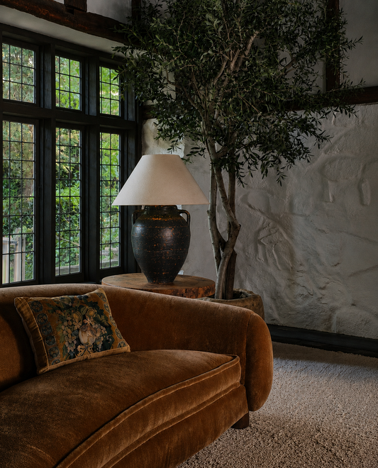 Large handcrafted ceramic table lamp with empire linen shade styled on a side table beside an olive tree in a characterful English country house interior.