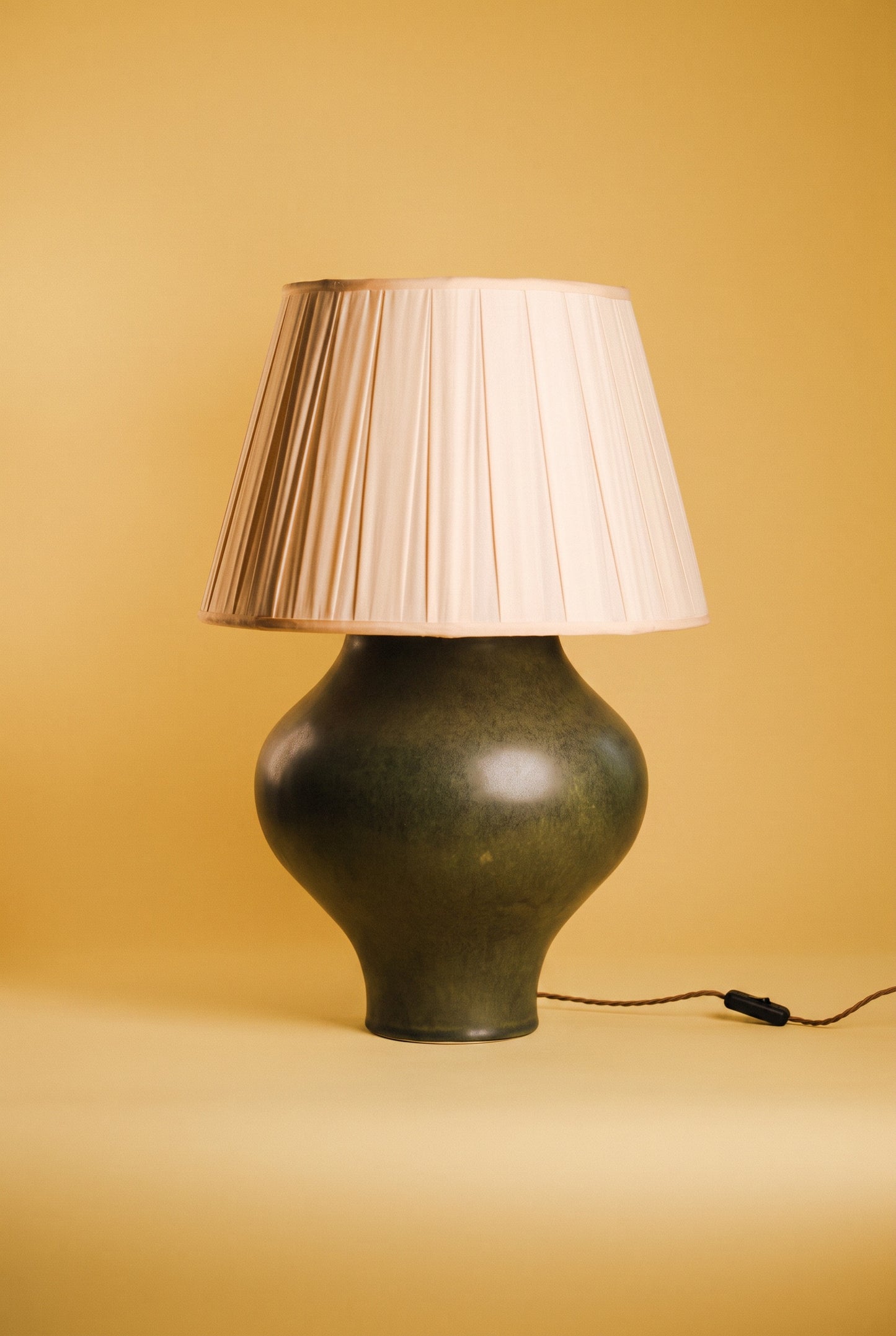 A vintage-style table lamp with a dark green glazed ceramic bulb and a pleated cream-colored fabric shade, set against a warm yellow background.